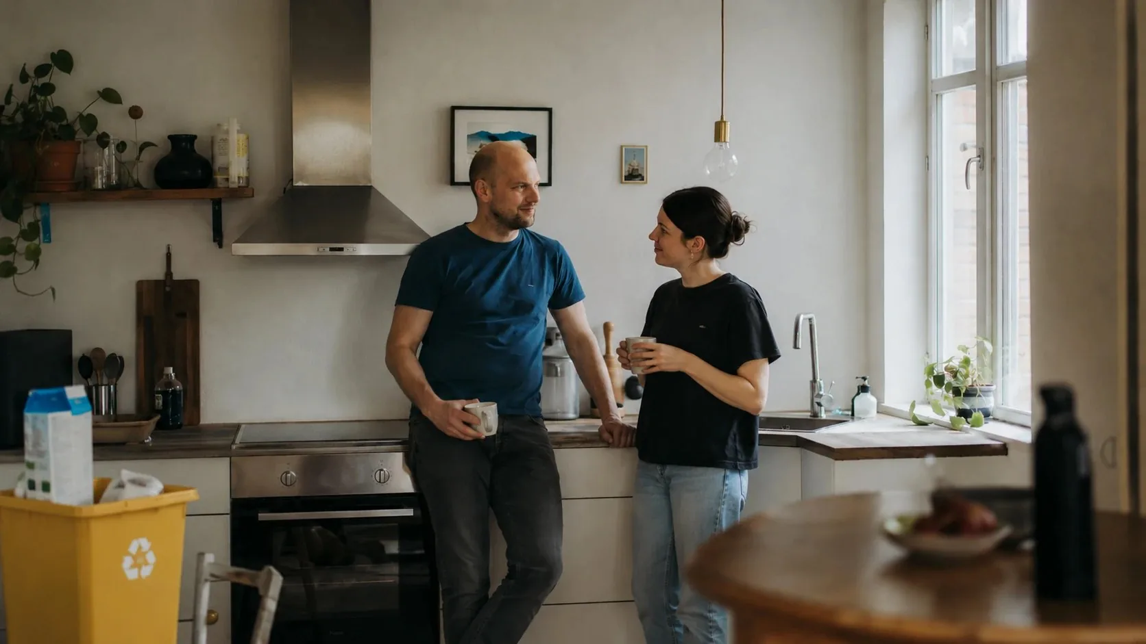 A couple standing together in their kitchen with coffee mugs in the morning; a milk carton and a yellow recycling bin in the corner